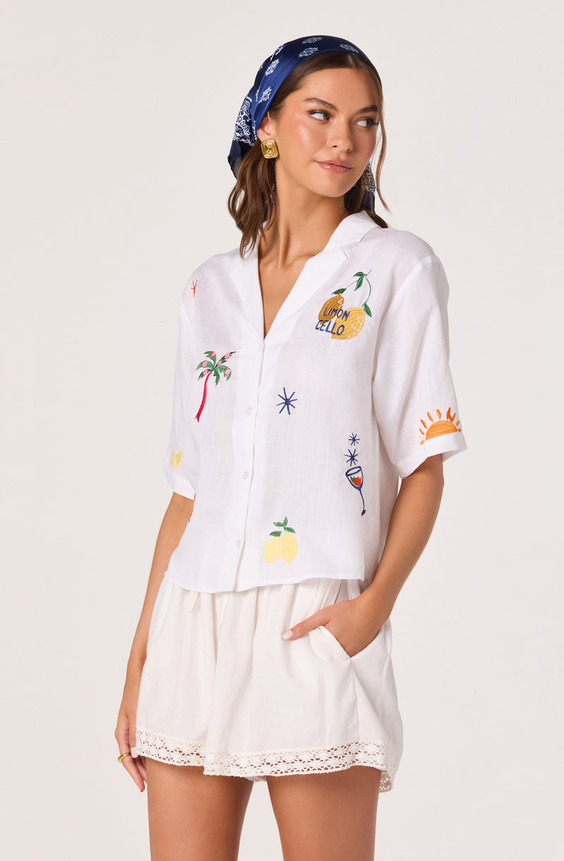 Woman wearing a white short-sleeve shirt with colorful embroidered summer-themed designs, paired with white shorts that have lace trim.
