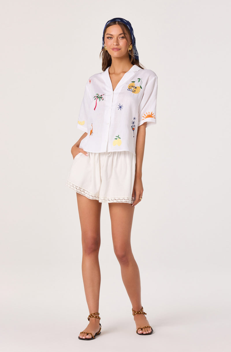 White short-sleeve shirt with colorful beach and summer-themed embroidery, paired with white shorts with lace trim.