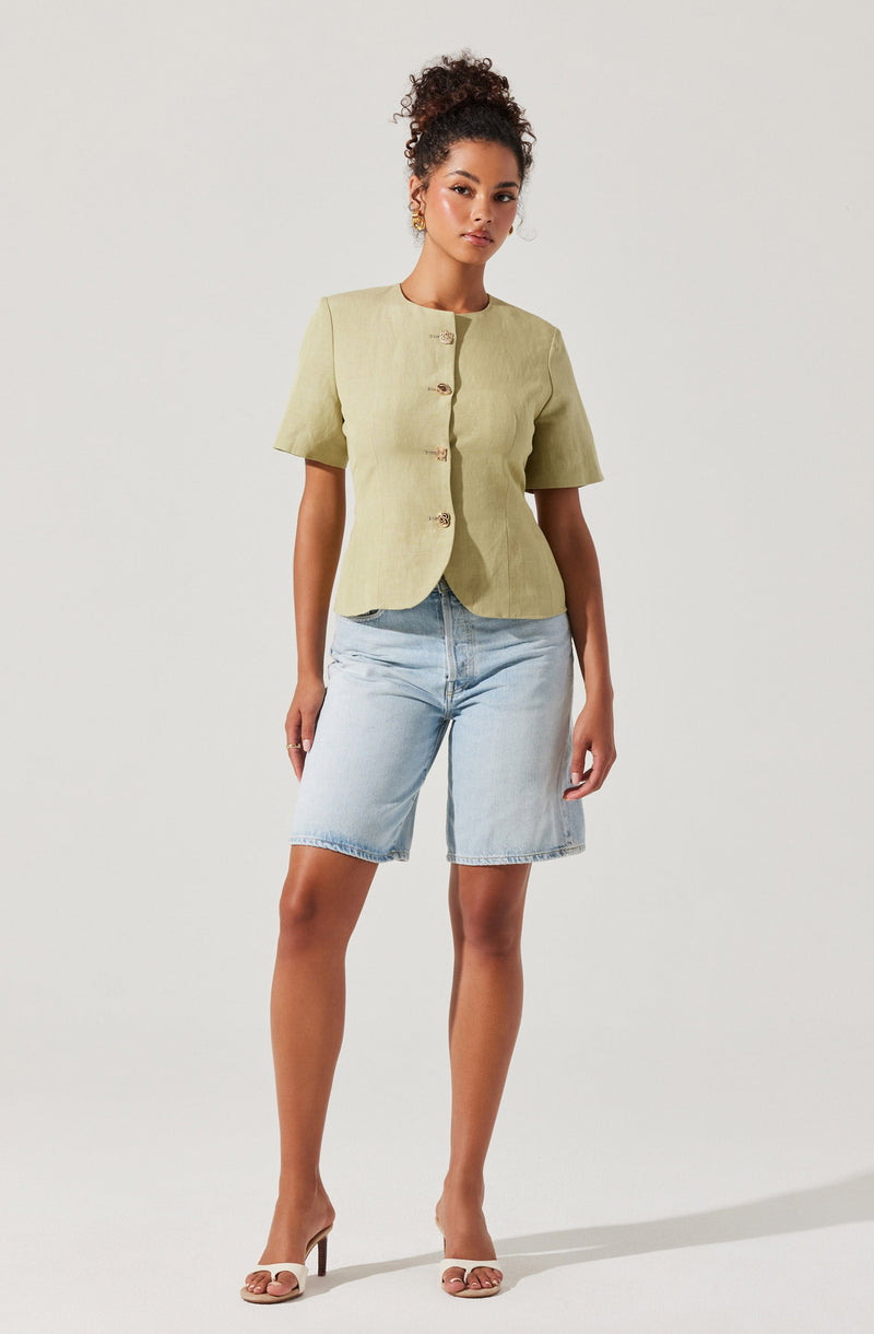 A woman is wearing a light green, short-sleeved, button-up jacket with a collarless neckline and three buttons down the front. She pairs the jacket with light blue denim knee-length shorts and beige open-toe high-heeled sandals.