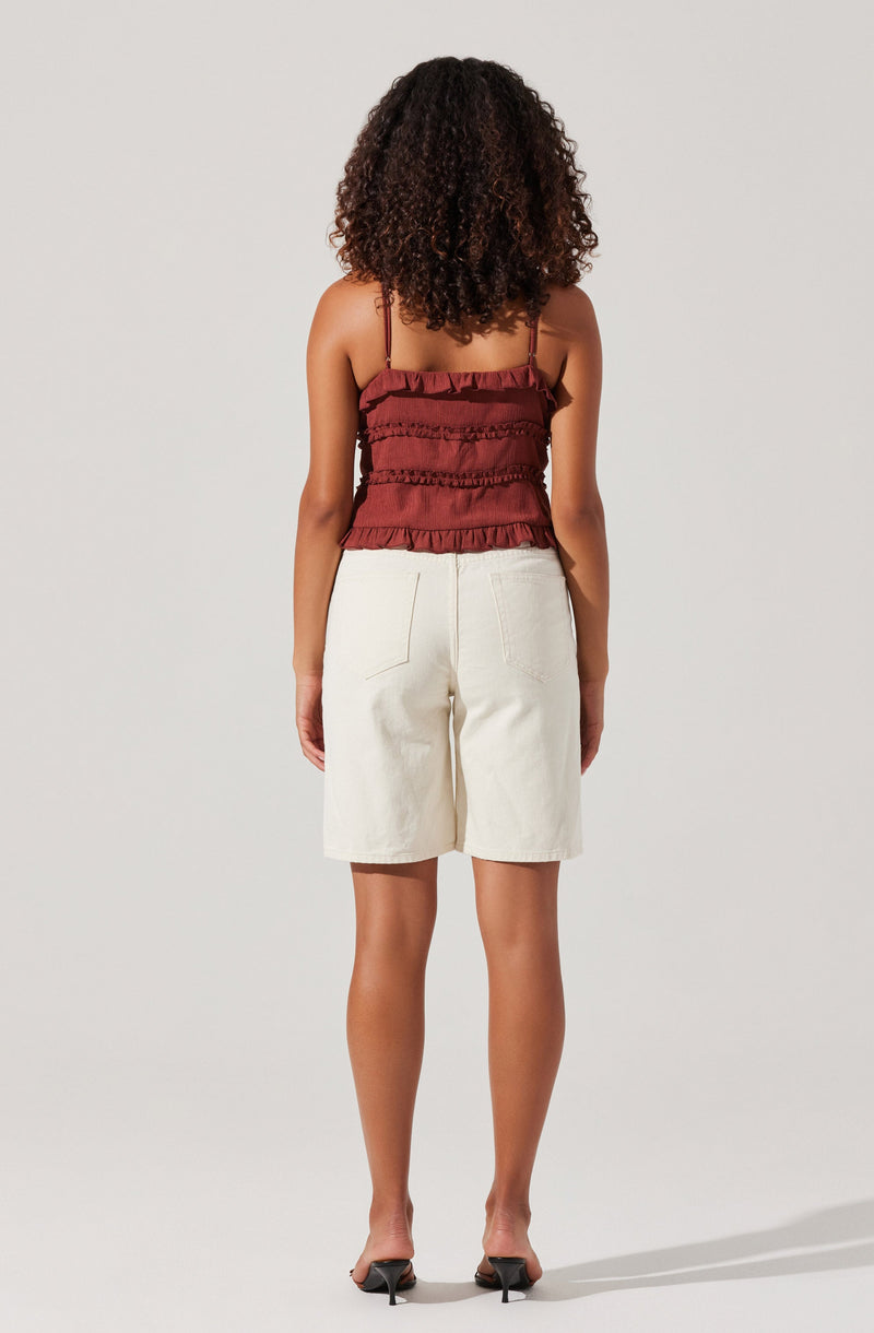 A woman is wearing a sleeveless, ruffled, reddish-brown top with thin spaghetti straps and horizontal ruffle details. She is also wearing high-waisted, loose-fitting cream-colored shorts with visible back pockets. The outfit is completed with black open-toe heeled sandals.
