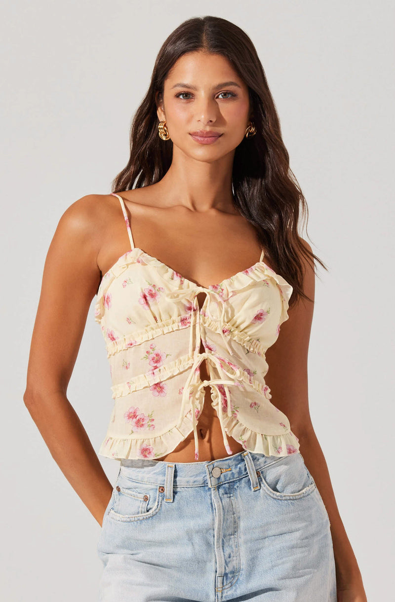 A woman is wearing a cream-colored, floral-patterned spaghetti strap top with ruffled edges and a front tie detail. She is pairing it with light-wash, high-waisted jeans.