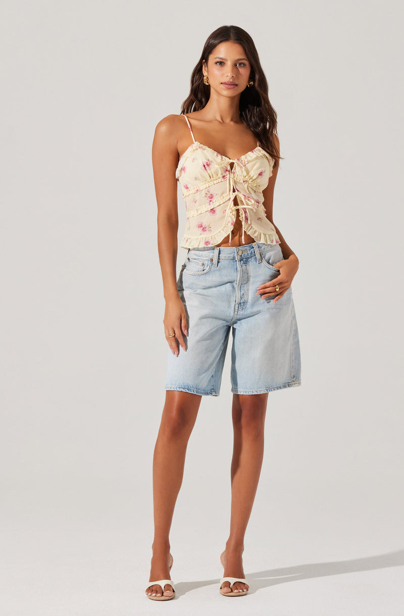 A woman is wearing a sleeveless, cream-colored top with pink floral patterns, featuring thin spaghetti straps, a ruched front with drawstrings, and ruffled edges. She is pairing it with light wash, knee-length denim shorts and white open-toe sandals.