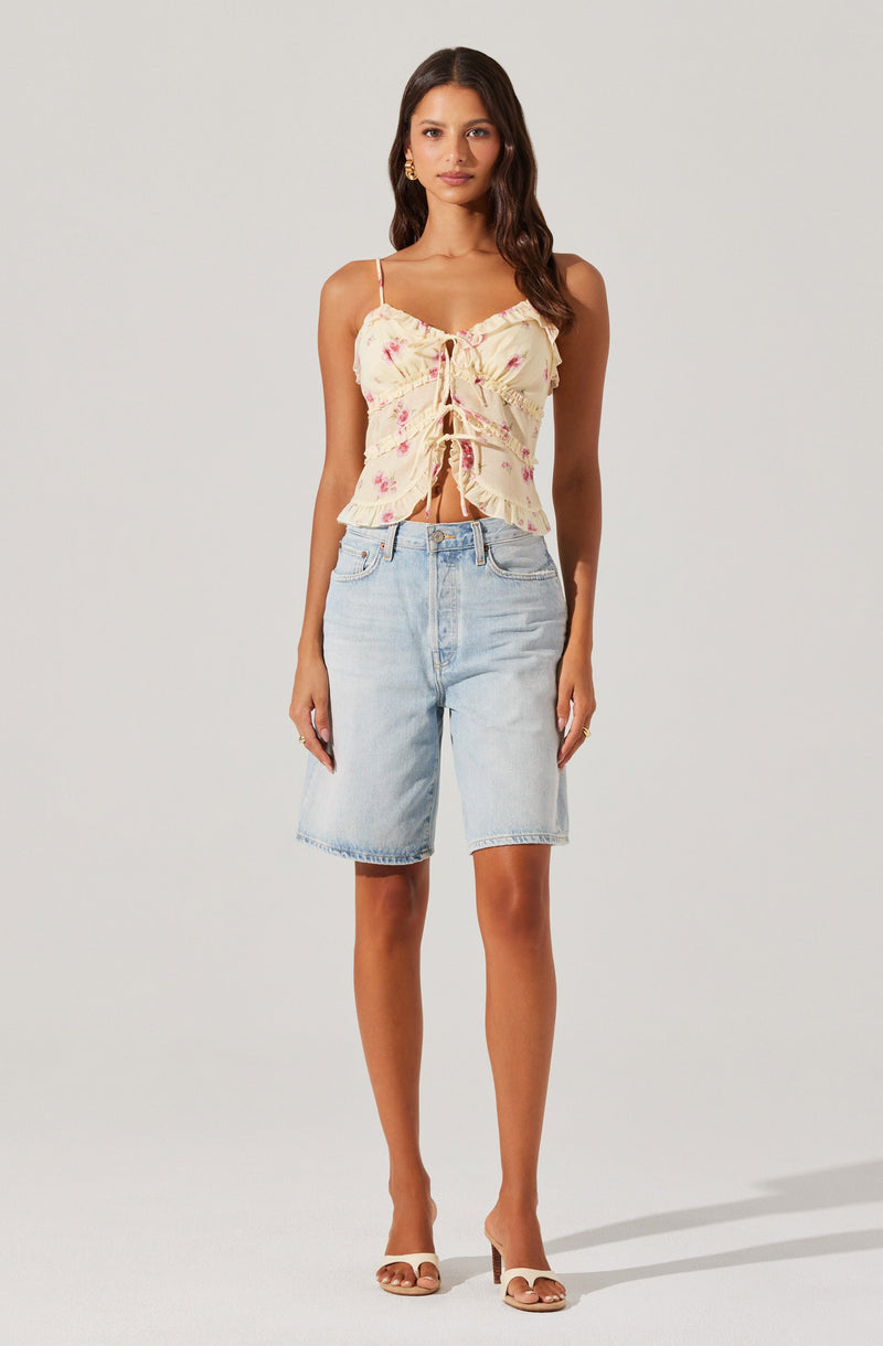 A woman is wearing a light-colored, sleeveless top with thin spaghetti straps and a tie-front detail, featuring a floral pattern in pink and yellow tones. She pairs the top with high-waisted, knee-length, light wash denim shorts and beige open-toe high heel sandals.