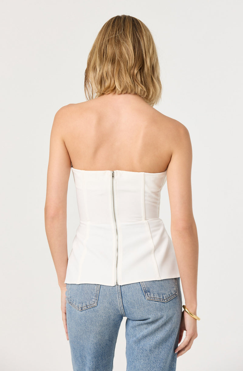 A woman is wearing a strapless, fitted, white top with vertical seam details and a back zipper. She is also wearing light blue high-waisted jeans and a gold bracelet on her left wrist.