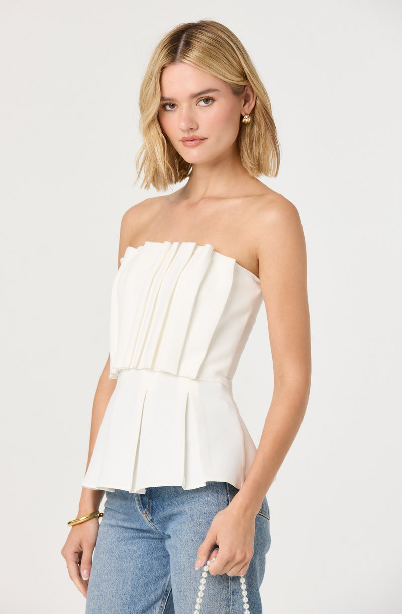 A woman is wearing a strapless white top with a pleated, ruffled front design and a fitted waist. She pairs it with light blue denim jeans. She accessorizes with small gold earrings and a gold bracelet.