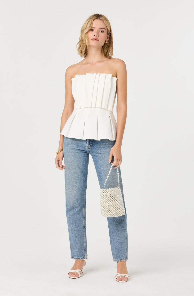 A woman is wearing a strapless white top with pleated vertical panels and a cinched waist, paired with light blue high-waisted jeans. She is holding a small, cream-colored woven handbag with a beaded strap and is wearing white open-toed heeled sandals.