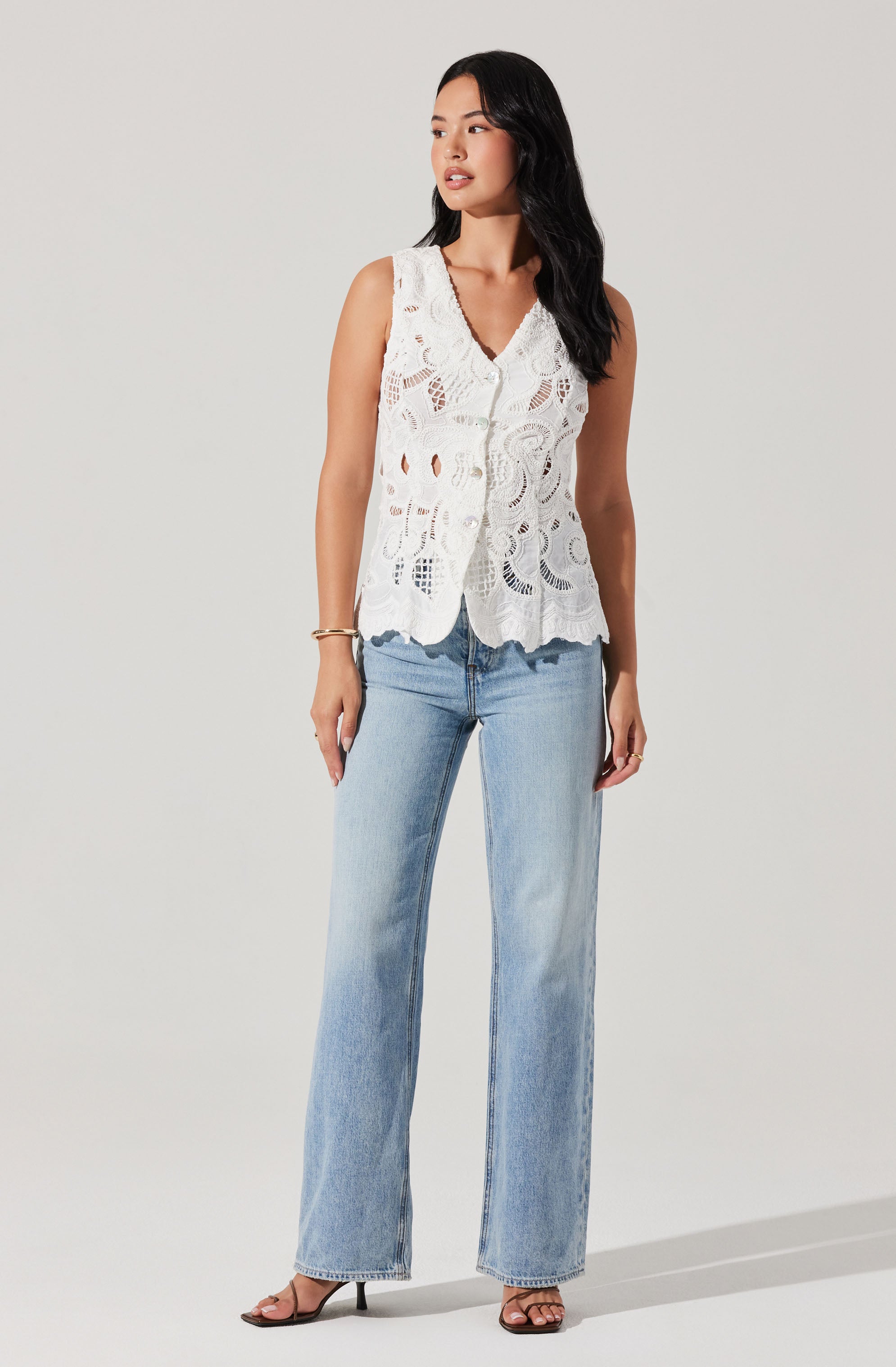 A woman is wearing a sleeveless, white, lace top with intricate cut-out patterns and scalloped edges. She pairs it with light-wash, wide-leg jeans and black high-heeled sandals.