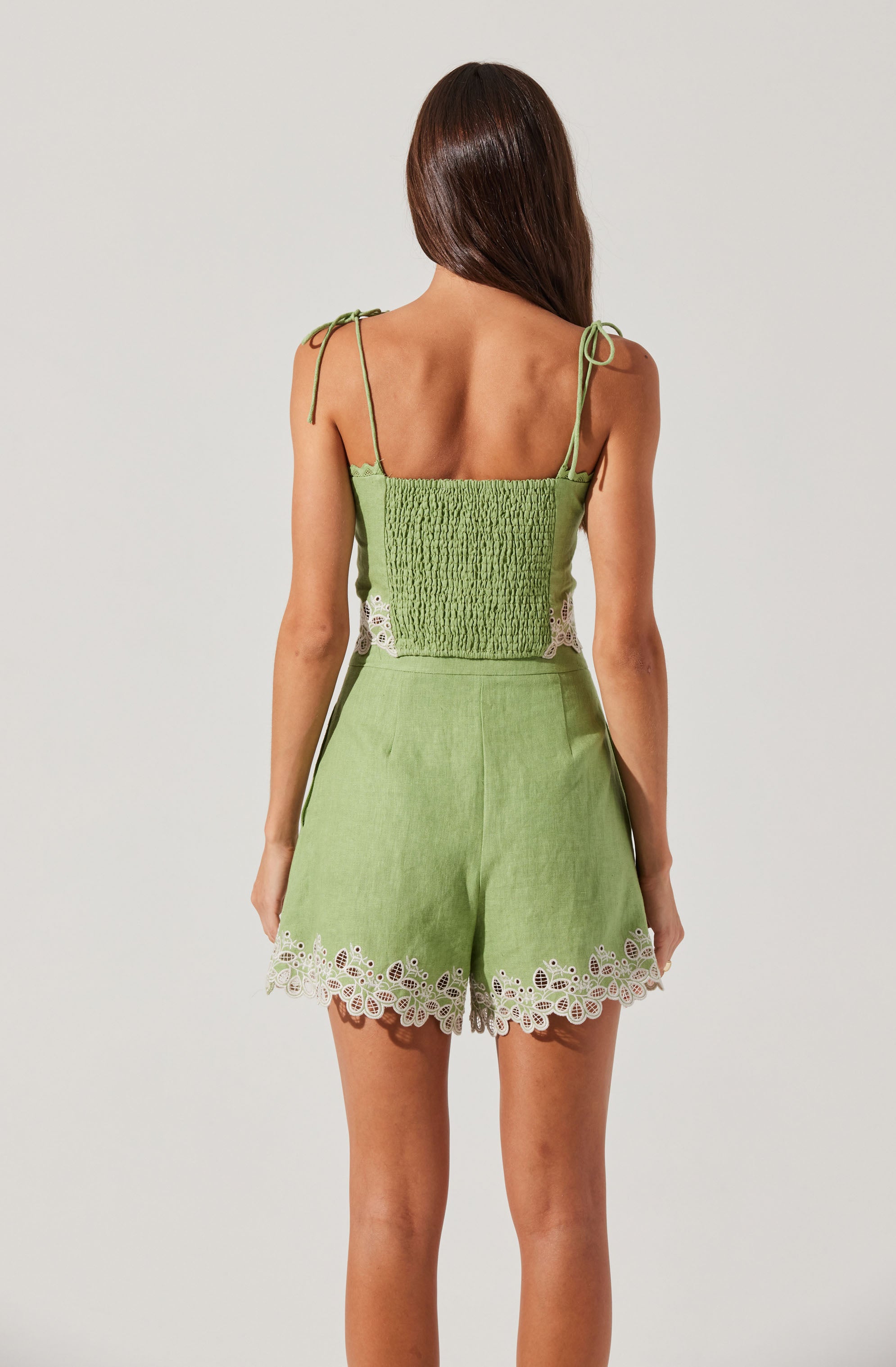 A woman is wearing a light green sleeveless top with spaghetti straps tied into bows on her shoulders. The top has a smocked, textured fabric in the front and small white lace panels on the sides. She is also wearing matching light green shorts with a decorative white lace scalloped trim along the hem.