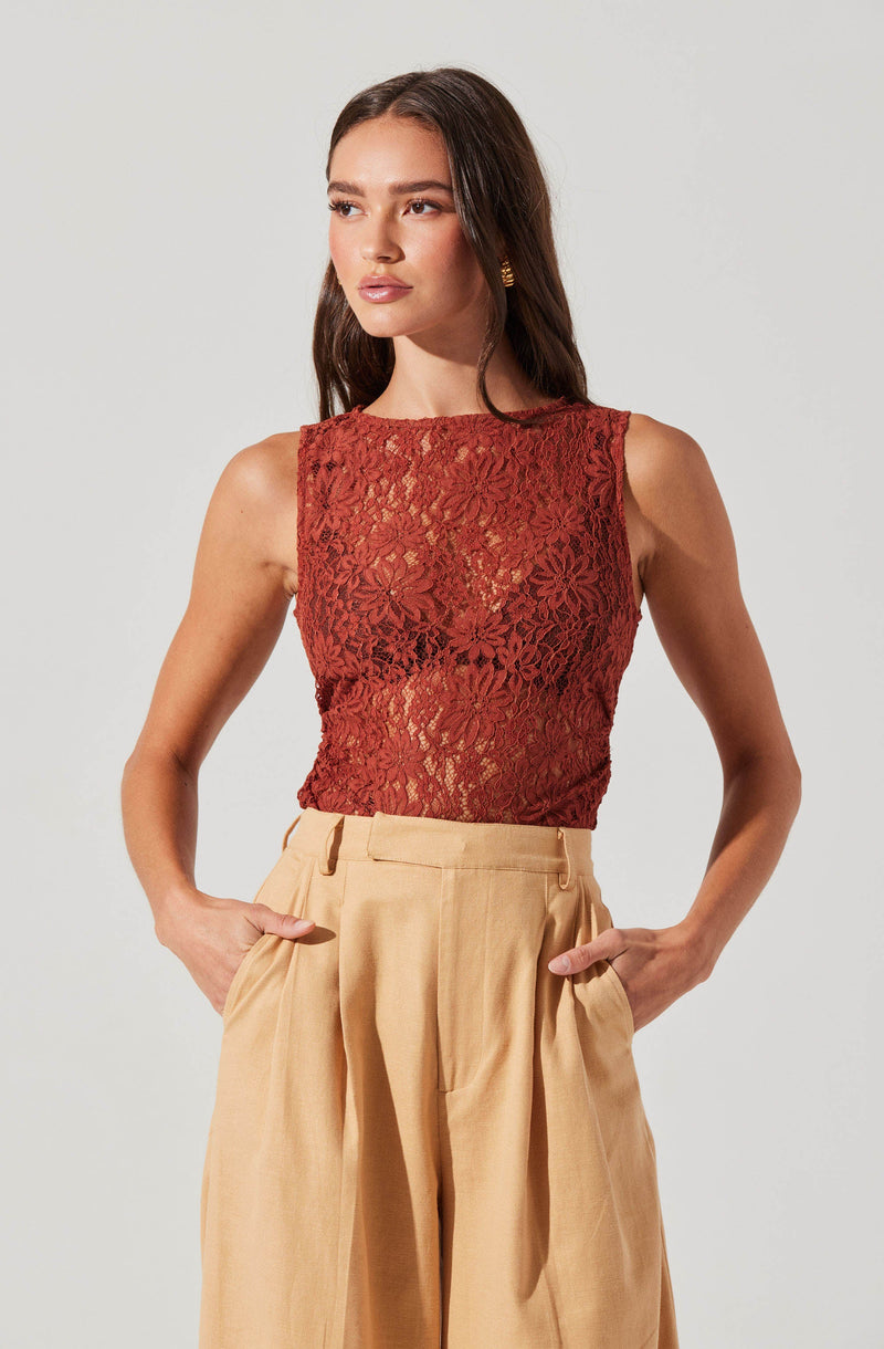 A woman is wearing a sleeveless, fitted top made of red lace with a floral pattern. The top is slightly sheer, revealing the skin underneath. She pairs it with high-waisted, wide-leg beige trousers that have pleats and side pockets.