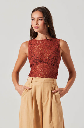 A woman is wearing a sleeveless, fitted top made of red lace with a floral pattern. The top is slightly sheer, revealing the skin underneath. She pairs it with high-waisted, wide-leg beige trousers that have pleats and side pockets.
