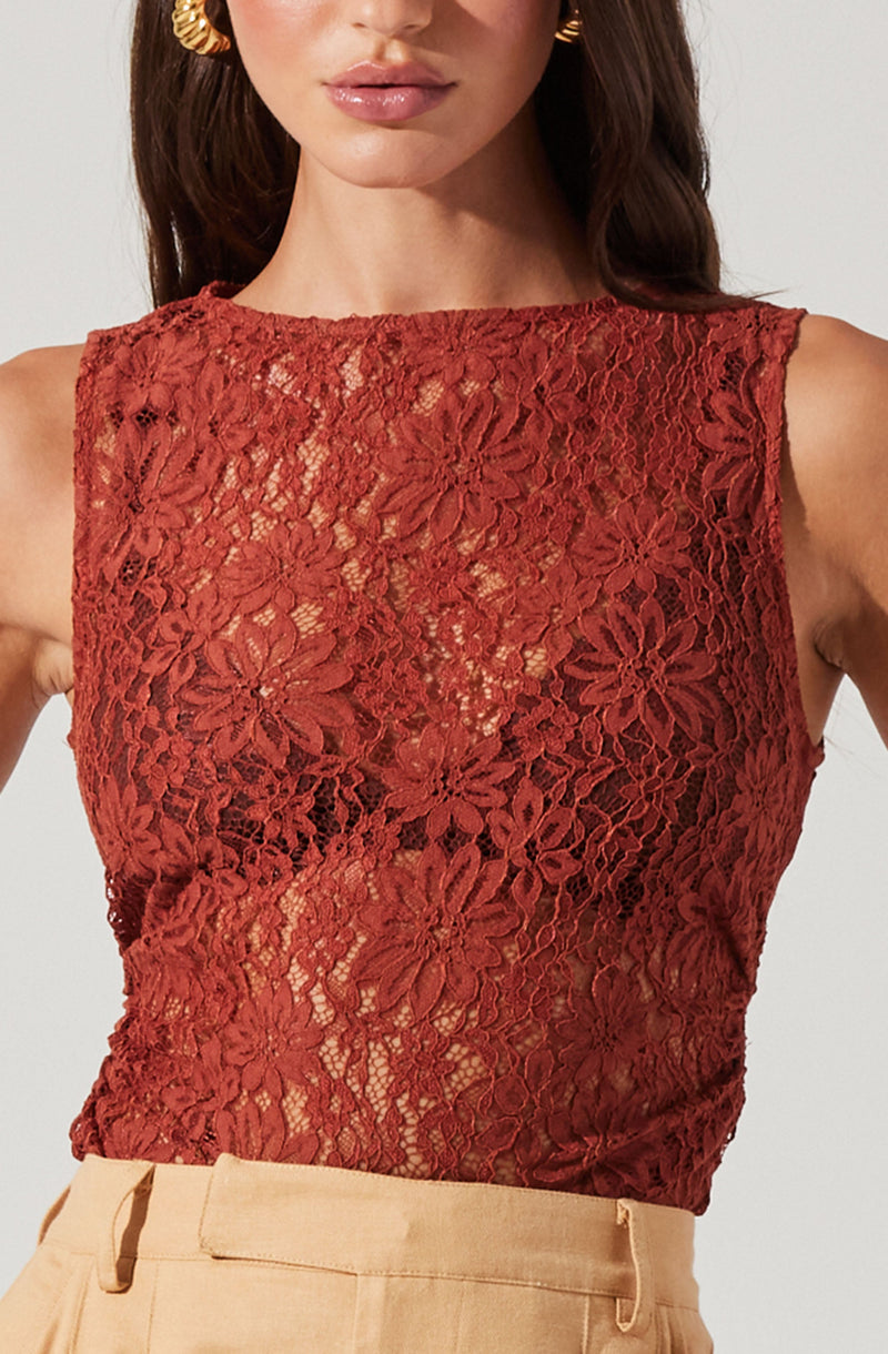 A woman is wearing a sleeveless, fitted top made of red lace fabric with a floral pattern. The lace is semi-sheer, revealing some skin beneath. She pairs it with high-waisted beige pants. She accessorizes with large, gold hoop earrings.