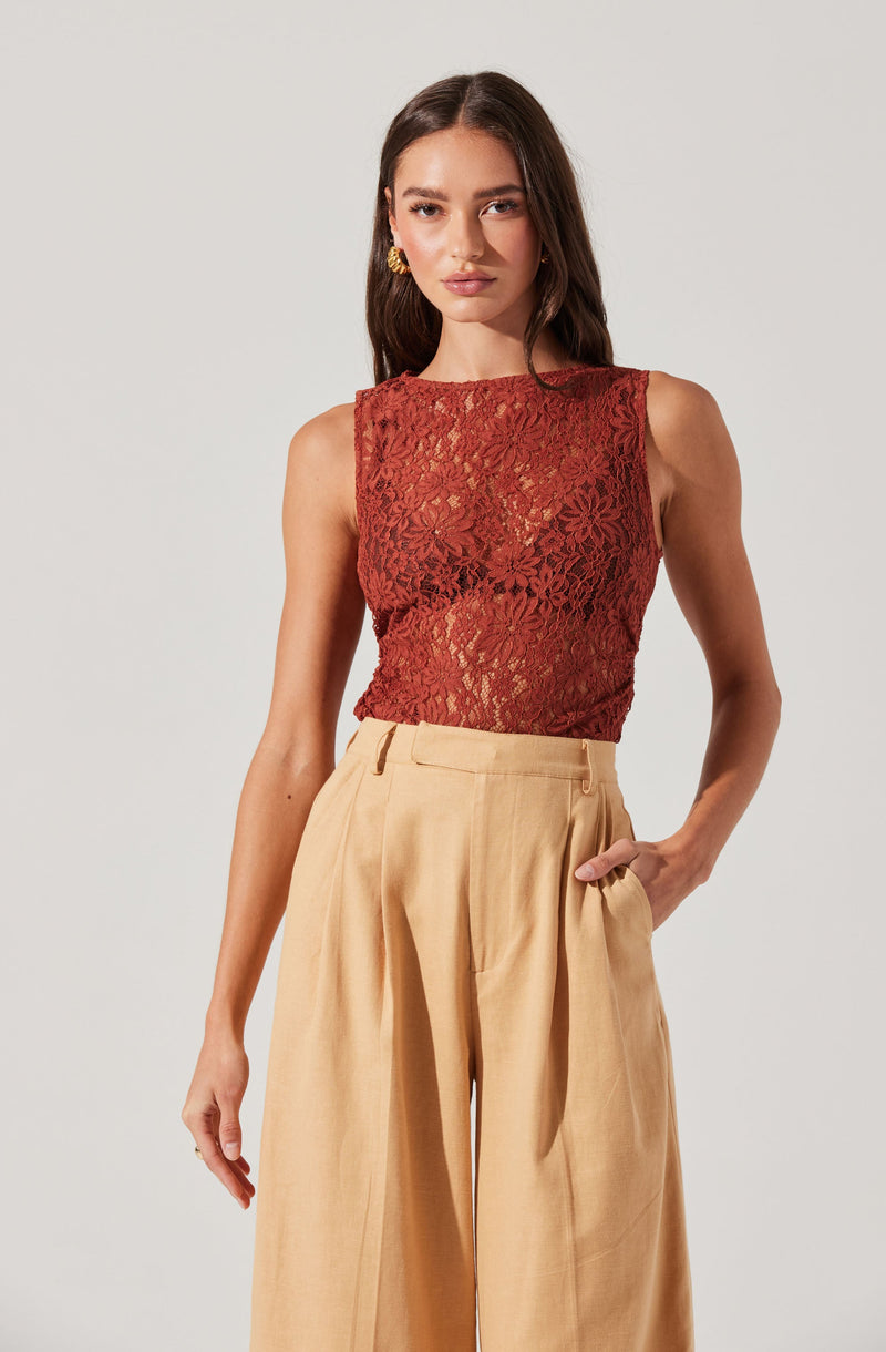 A woman is wearing a sleeveless, rust-colored lace top with floral patterns, paired with high-waisted, beige wide-leg trousers.