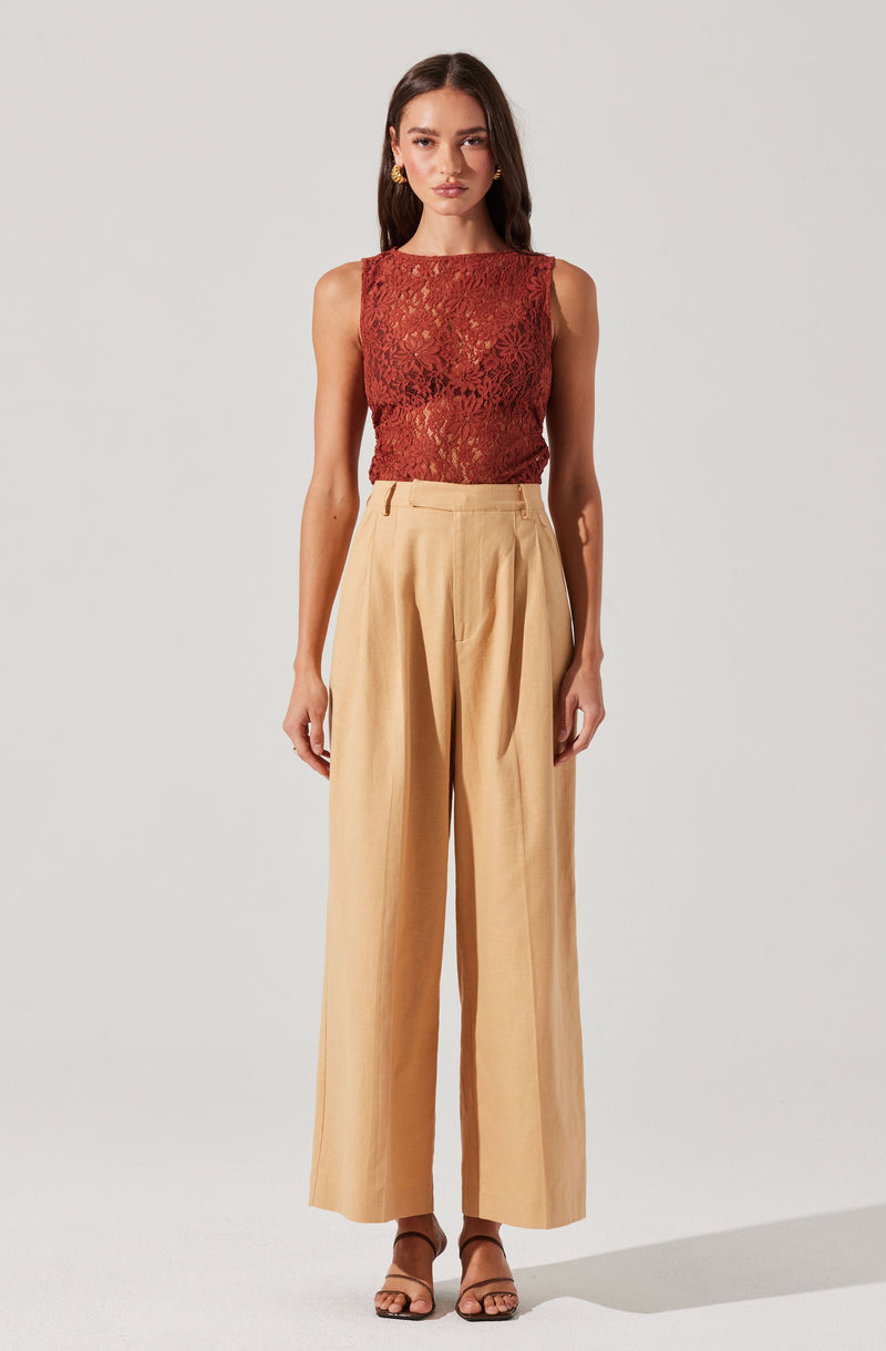 A woman is wearing a sleeveless, fitted, red lace top with floral patterns paired with high-waisted, wide-leg beige trousers that have pleats at the front. She is also wearing open-toe sandals.