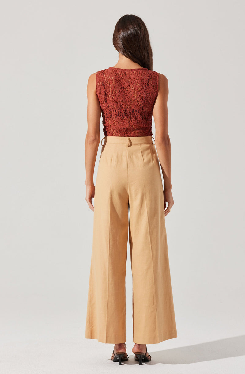 A woman is wearing a sleeveless, fitted top made of red lace fabric with floral patterns. She pairs it with high-waisted, wide-leg beige trousers that have pleats at the waist. She is also wearing black open-toe heels.