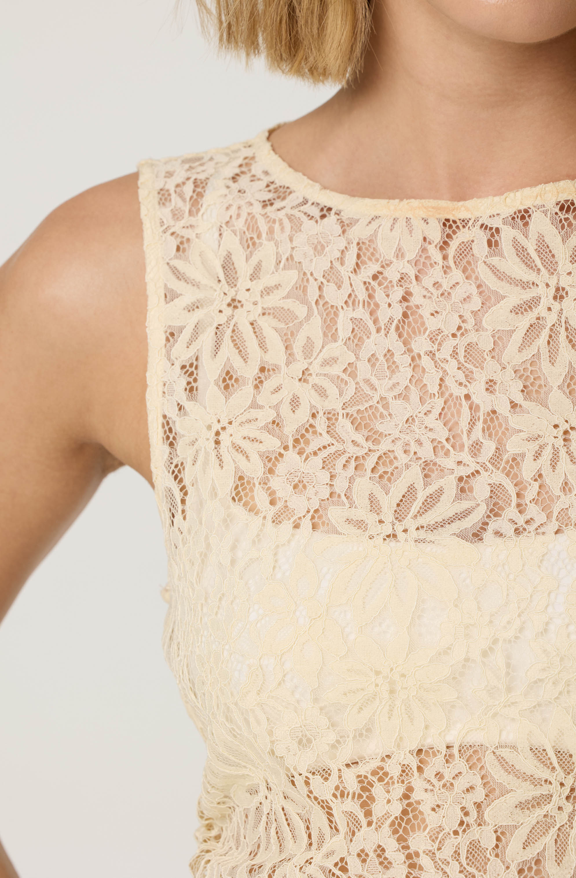 A woman is wearing a sleeveless, cream-colored lace top featuring intricate floral lace patterns with a high, rounded neckline. The lace fabric is semi-transparent, and the floral design covers the upper chest and shoulders, with a more solid lace section around the waist.