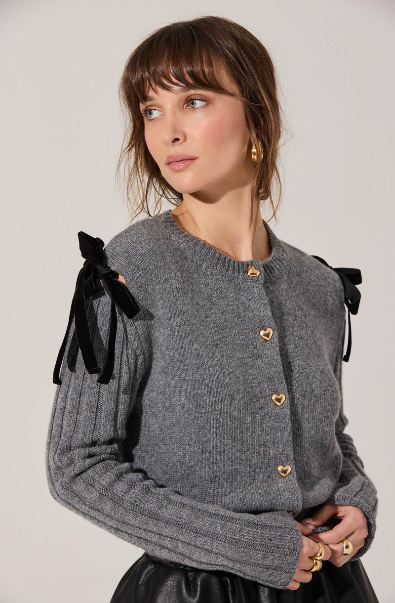 A woman is wearing a gray knitted cardigan with gold heart-shaped buttons down the front. The cardigan features black ribbon bows on the shoulders. She is also wearing a black leather skirt and gold jewelry, including hoop earrings and rings.