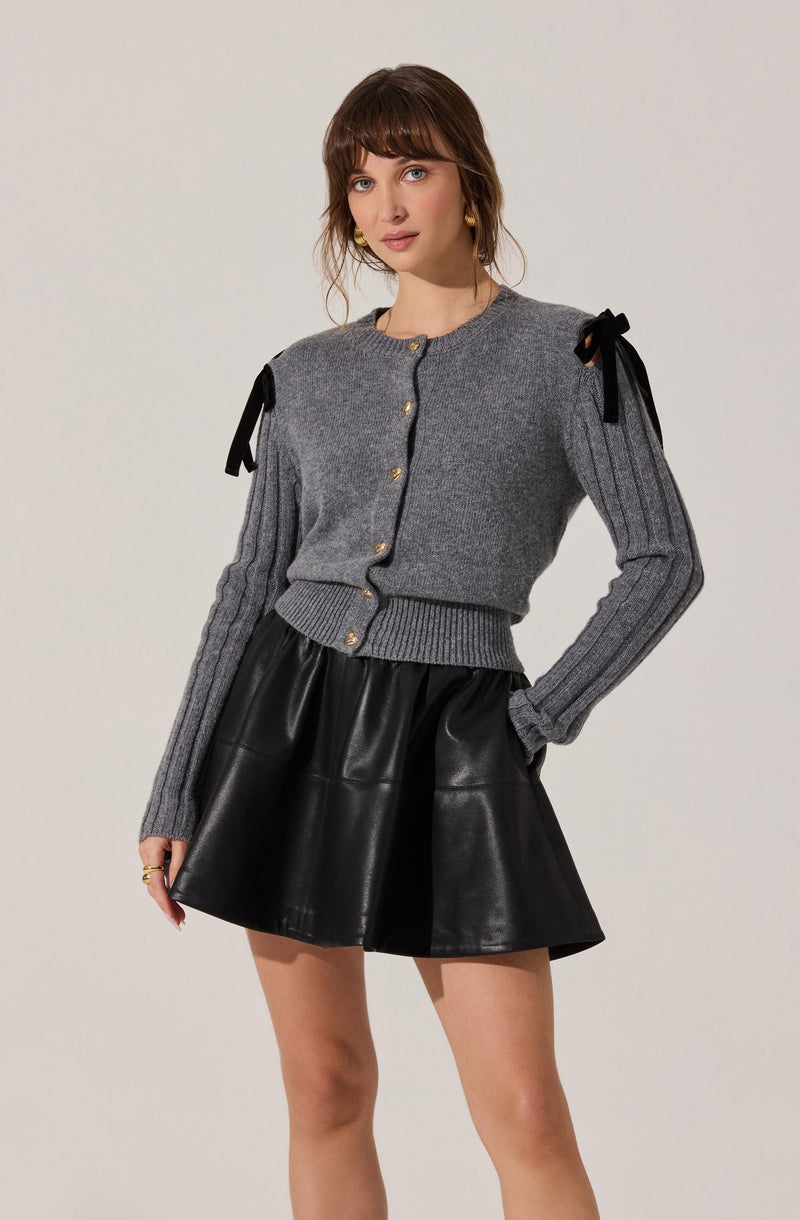 A woman is wearing a gray knit sweater with black bows on the shoulders and gold buttons down the front. She is also wearing a black leather mini skirt.