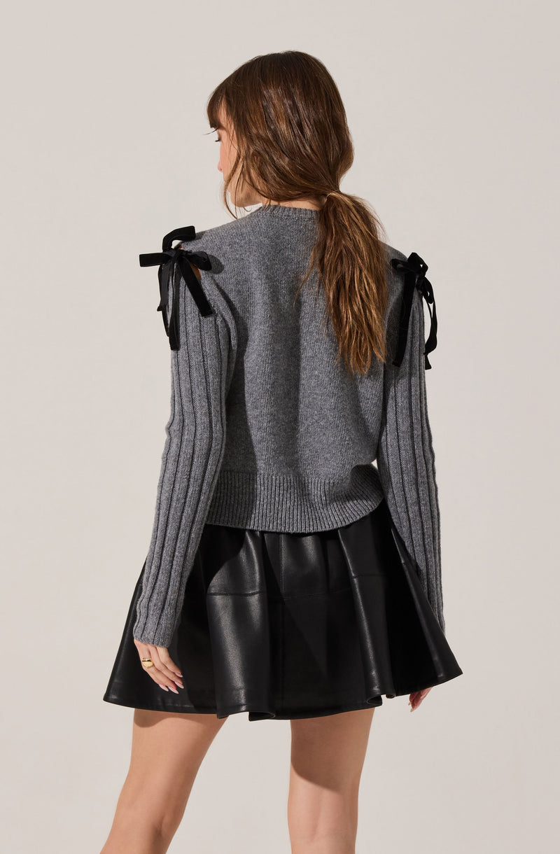 A woman is wearing a gray knit sweater with ribbed details on the sleeves and shoulder bows made of black ribbon. She pairs the sweater with a black, shiny, leather-like skirt.
