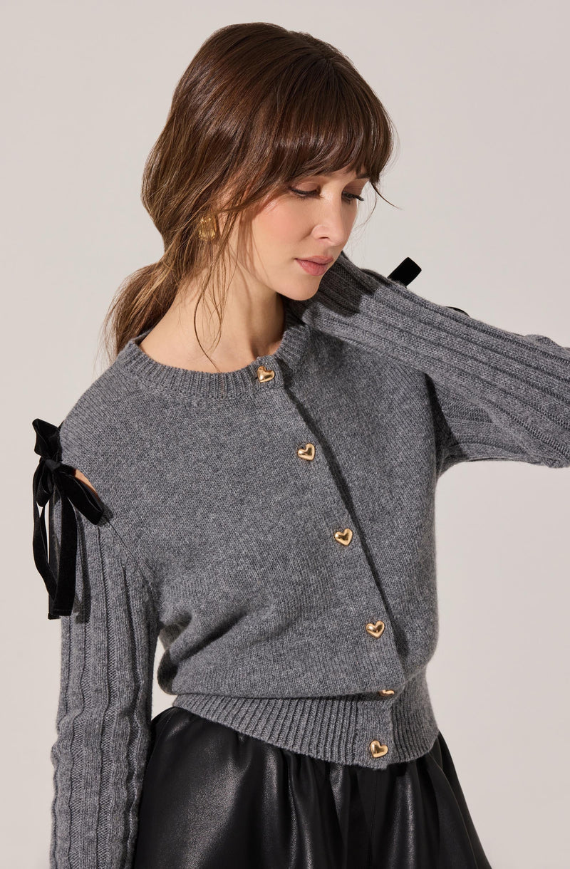 A woman is wearing a grey knit cardigan with gold heart-shaped buttons down the front. The cardigan has ribbed cuffs and hem, and features black ribbon ties on the shoulders. She is also wearing a black skirt made of a shiny, leather-like material.