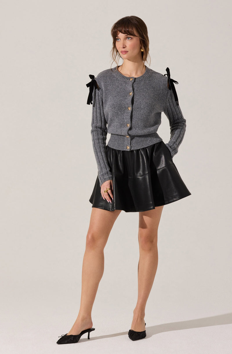 A woman is wearing a gray cardigan with black ribbon bows on the shoulders, gold buttons down the front, and ribbed cuffs and hem. She pairs it with a black, shiny, flared skirt and black pointed-toe heels with small bows.