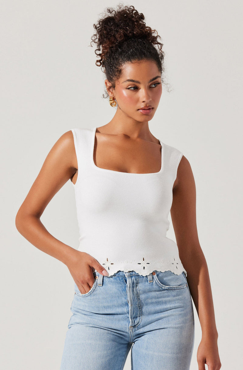 A woman is wearing a sleeveless white top with a square neckline and decorative cut-out flower patterns along the hemline, paired with light blue denim jeans.