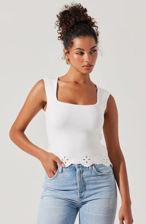 A woman is wearing a sleeveless white top with a square neckline and decorative cut-out flower patterns along the hemline, paired with light blue denim jeans.