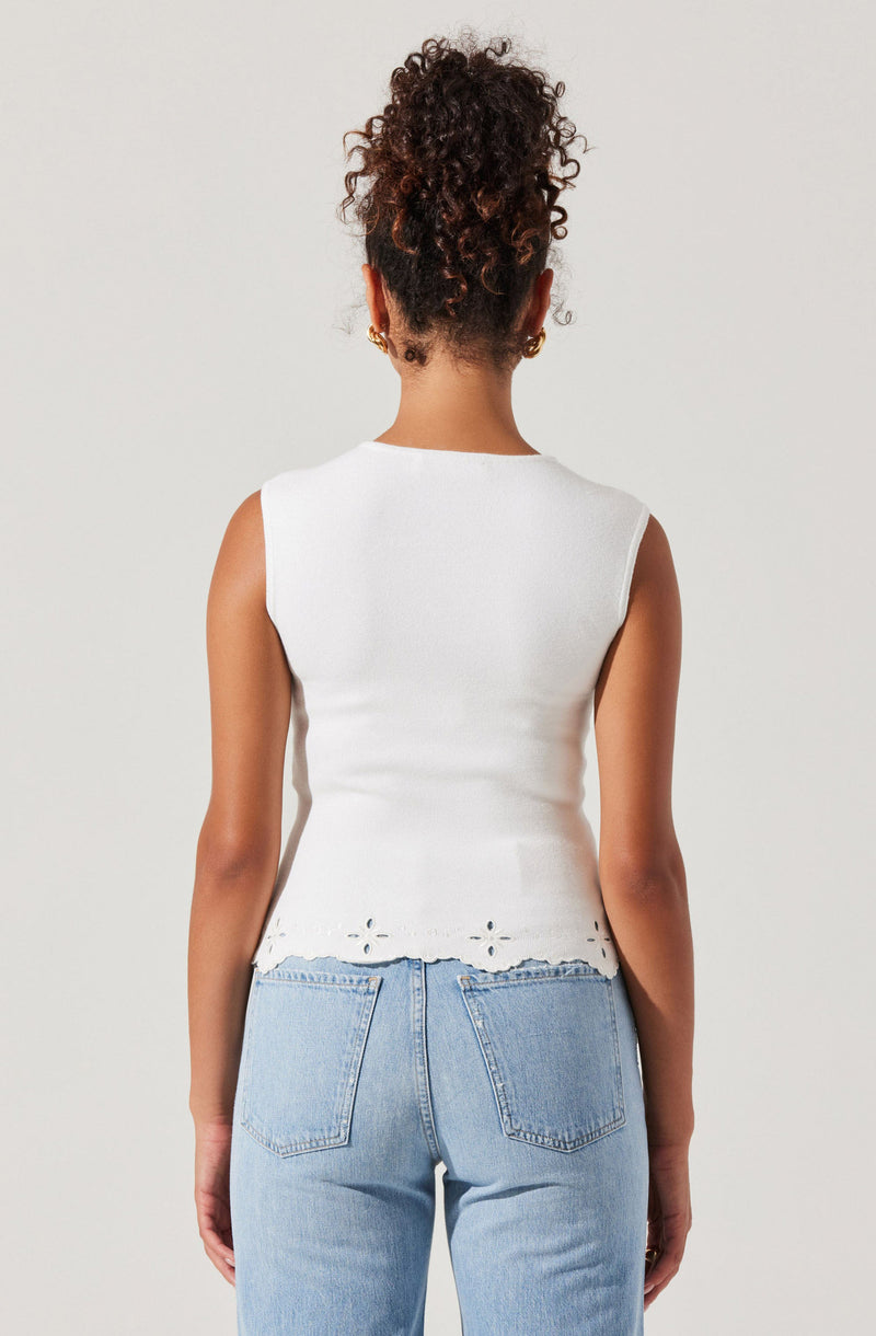 A woman is wearing a sleeveless white top with a scalloped hem and eyelet detailing at the bottom. She is also wearing light blue high-waisted jeans.