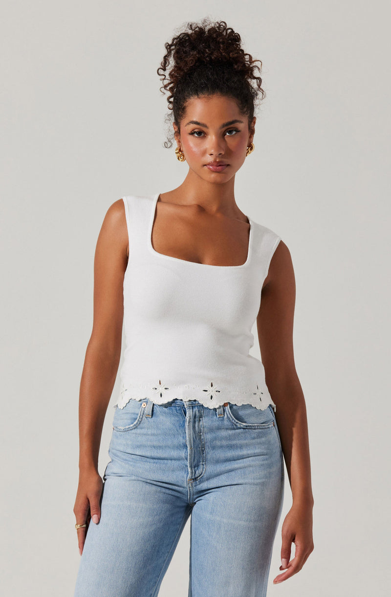 A woman is wearing a sleeveless white top with a square neckline and embroidered eyelet details along the hemline, paired with light-wash high-waisted jeans.