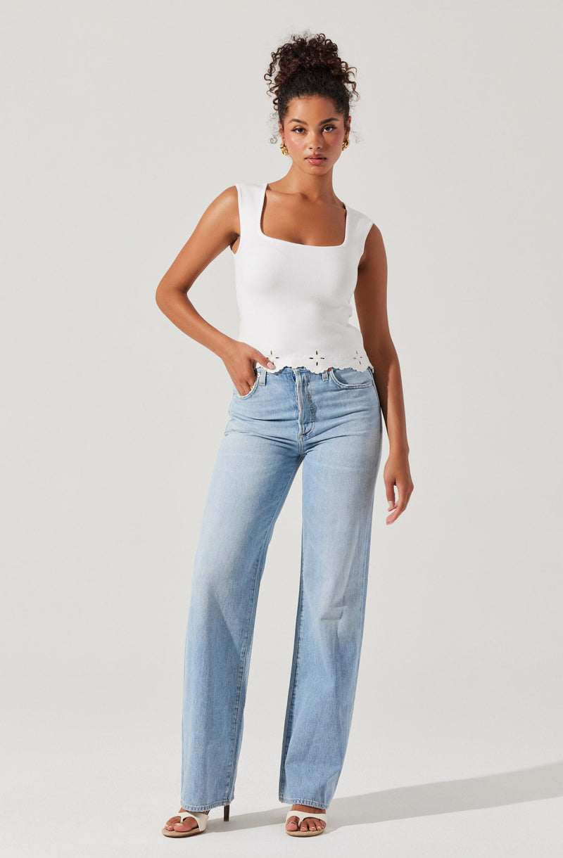 A woman is wearing a sleeveless white top with a square neckline and a scalloped hem, paired with light-wash, high-waisted, straight-leg jeans. She is also wearing open-toe sandals with a block heel.