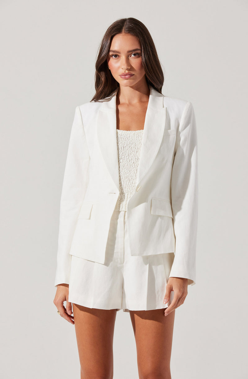 A woman is wearing a white tailored blazer with a single-button closure, structured shoulders, and flap pockets. She pairs it with matching high-waisted white shorts that have a loose fit and pleats. Underneath, she has on a cream-colored, textured sleeveless top.