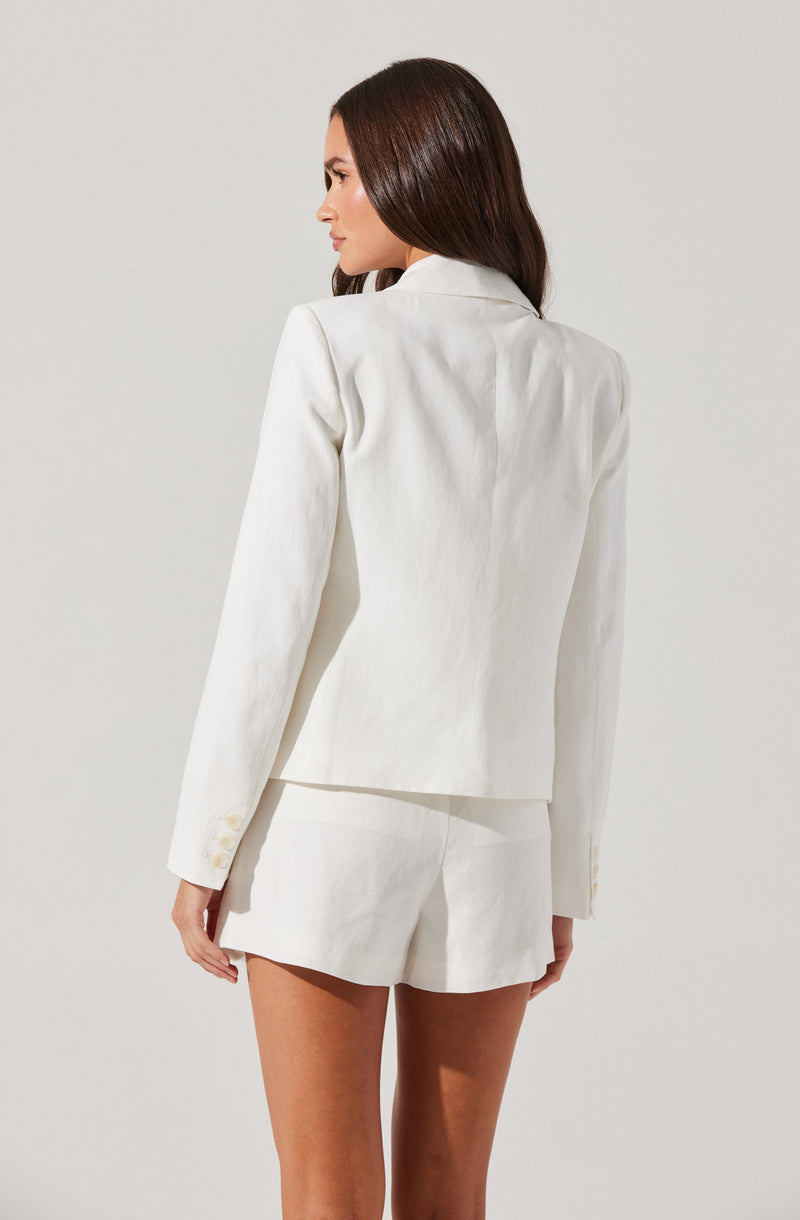 A woman is wearing an off-white, tailored blazer with long sleeves and a pair of matching off-white shorts. The blazer has a structured fit with subtle shoulder padding and buttons on the sleeve cuffs. The shorts are high-waisted with a clean, minimal design.