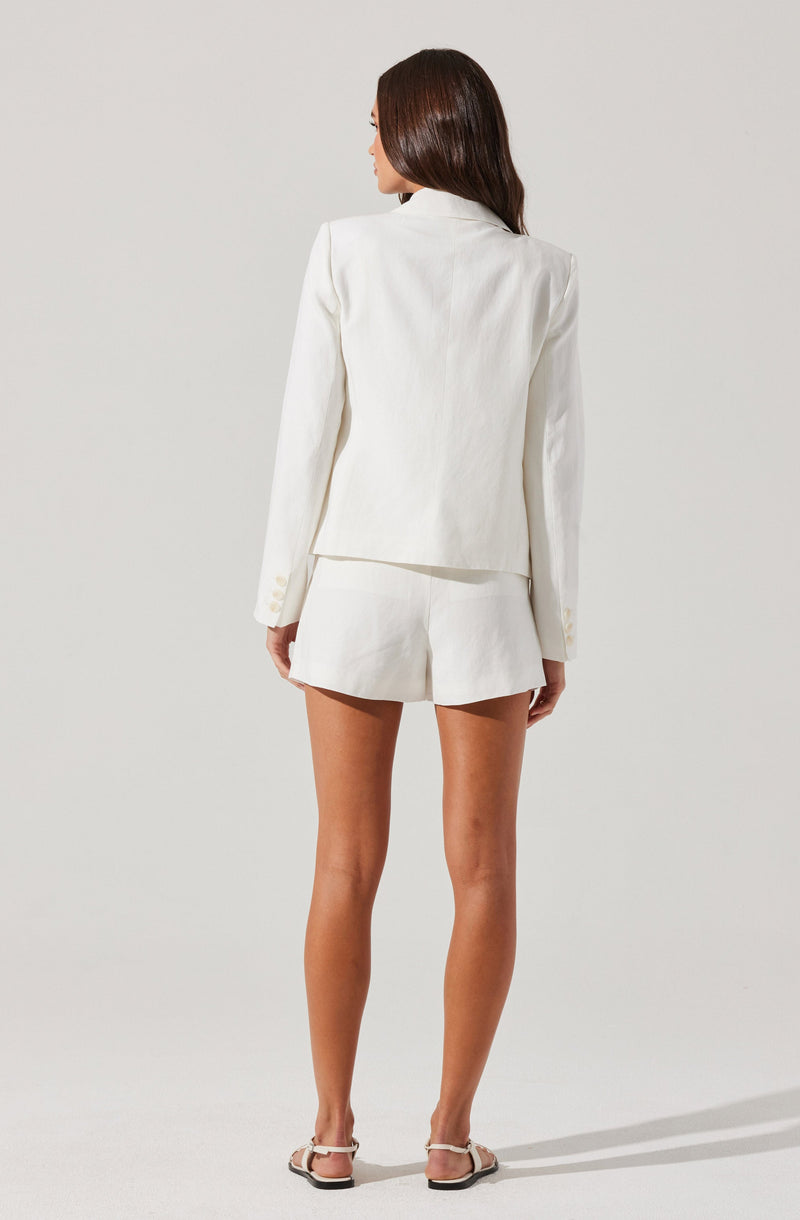 A woman is wearing an all-white outfit consisting of a tailored blazer with long sleeves and a notched collar, paired with matching high-waisted shorts. She has long, straight brown hair and is wearing flat sandals with ankle straps.