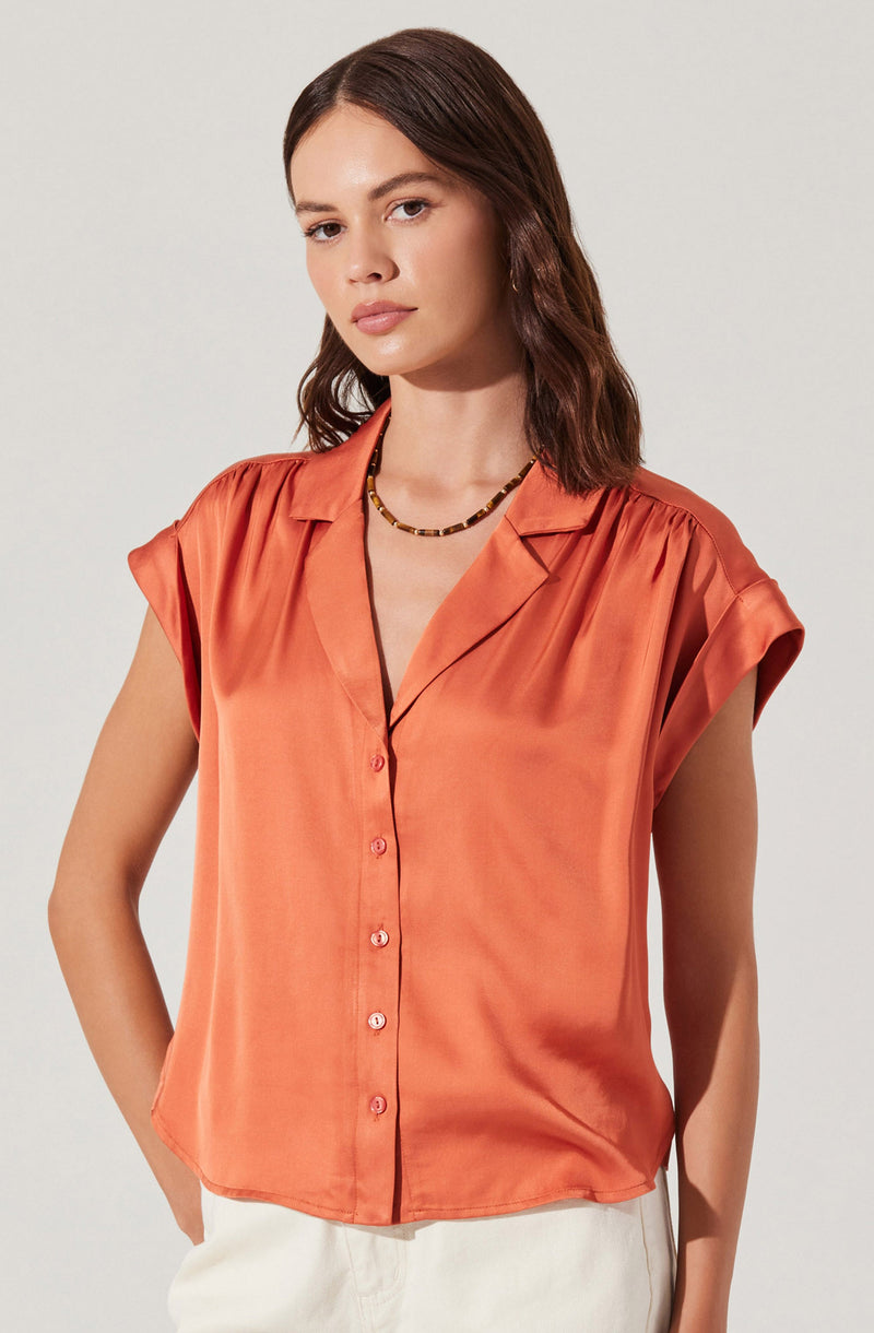 A woman is wearing a short-sleeved, button-up satin blouse in a warm, orange hue. The blouse features a notched collar and slightly puffed shoulders. She pairs it with a beaded necklace with a mix of dark and light beads.
