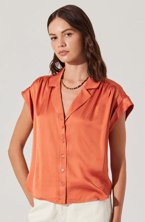 A woman is wearing a short-sleeved, button-up satin blouse in a warm, orange hue. The blouse features a notched collar and slightly puffed shoulders. She pairs it with a beaded necklace with a mix of dark and light beads.