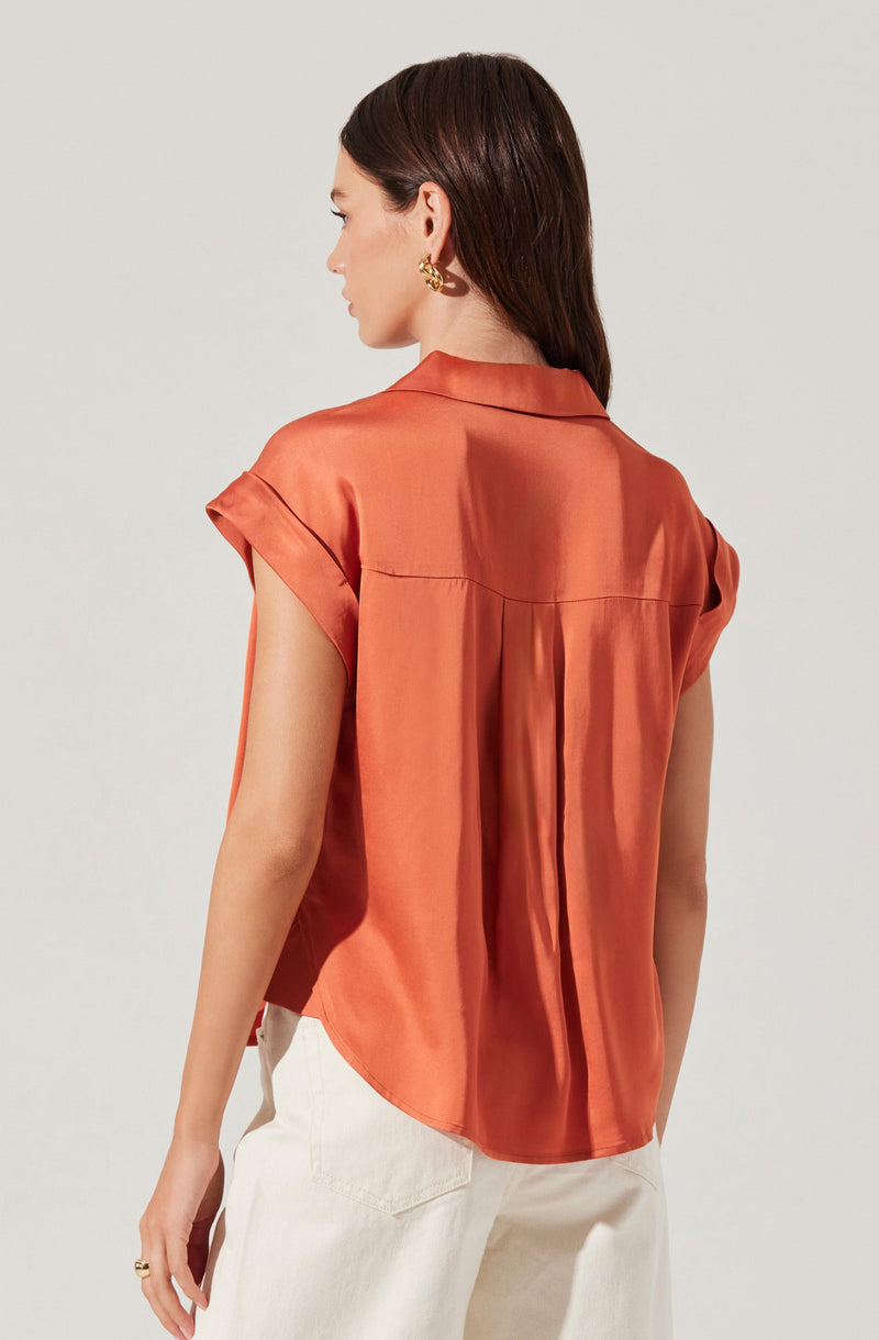 A woman is wearing a short-sleeved, rust-colored silk or satin blouse with a loose fit. The blouse features a collar and pleats down the back. She is also wearing off-white or cream-colored pants.