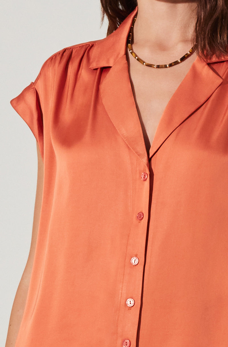 A woman is wearing a sleeveless, button-up blouse made of shiny, satin-like fabric in a coral-orange color. The blouse has a classic collar and features small, round buttons. She is also wearing a beaded necklace with dark and light brown beads.