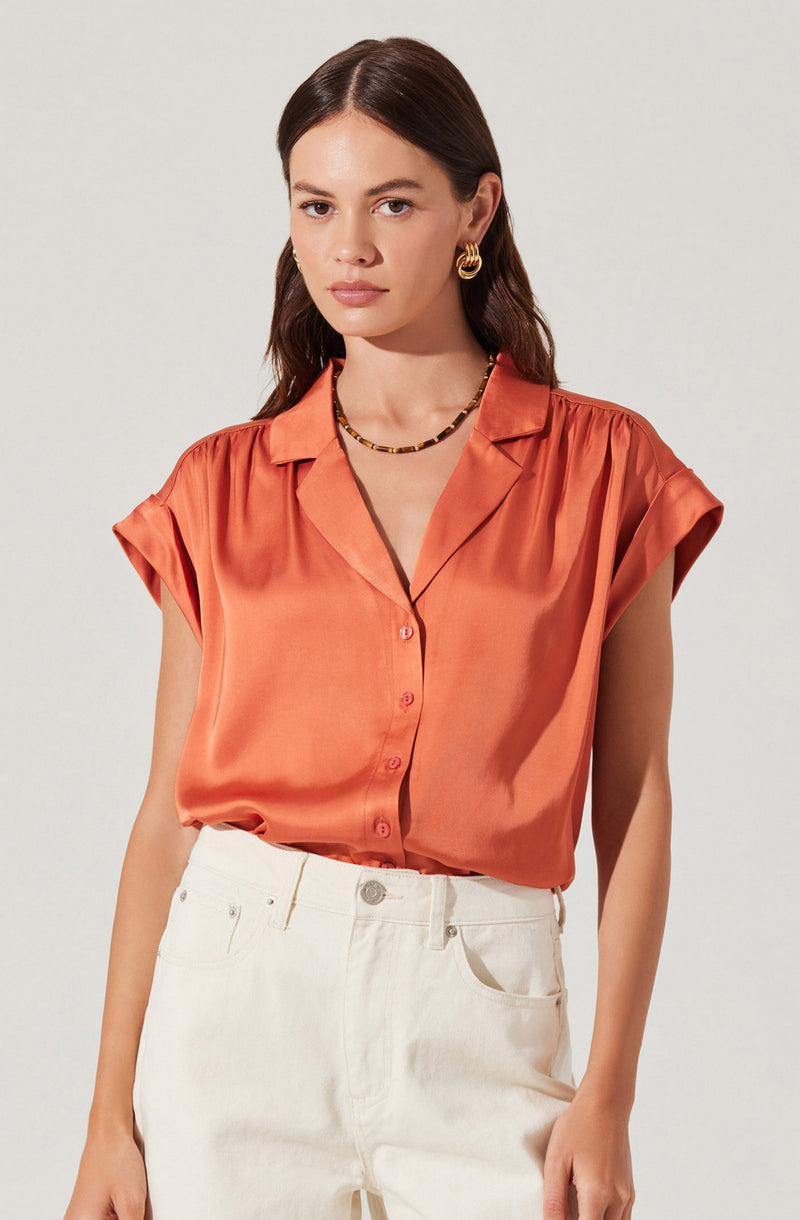 A woman is wearing a short-sleeved, collared, button-up blouse made of a shiny, satin-like fabric in a warm, coral-orange color. The blouse features rolled-up sleeves and a relaxed fit. She pairs it with high-waisted white jeans. She accessorizes with gold hoop earrings and a beaded necklace.