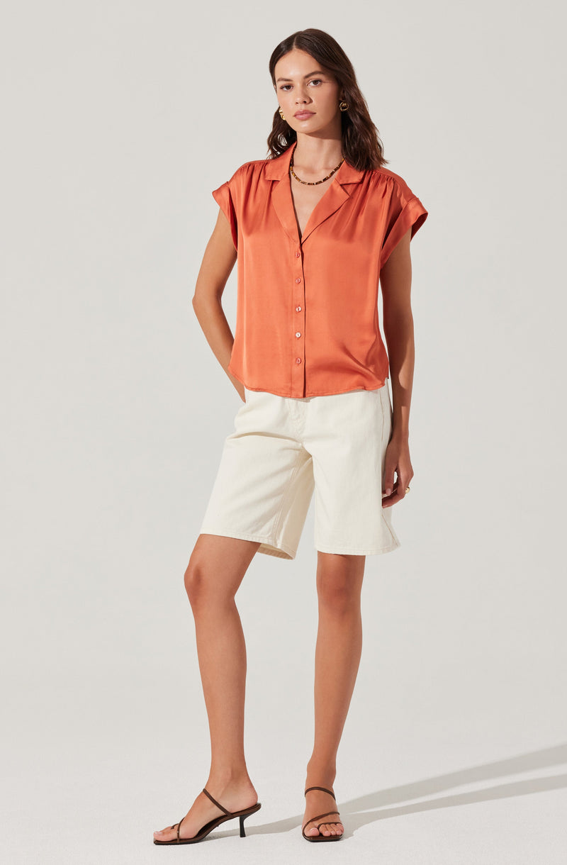 A woman is wearing a sleeveless, orange satin button-up blouse with a collar, paired with high-waisted, off-white knee-length shorts. She has on black strappy high-heeled sandals and accessorizes with earrings and a necklace.