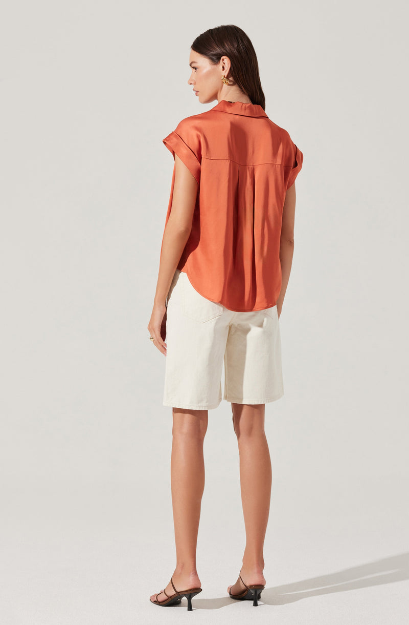 A woman is wearing a loose, short-sleeved, rust-colored satin blouse with a back pleat and a collar. She pairs it with knee-length, white linen shorts and black open-toe sandals with a small heel.