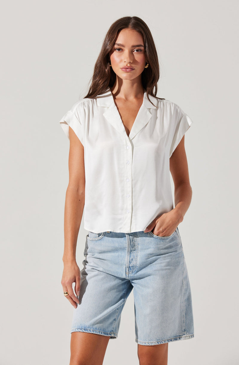 A woman wearing a white, short-sleeved, collared button-up shirt with a slightly loose fit. She pairs it with light blue, high-waisted denim shorts that have a relaxed fit and reach just above the knees.