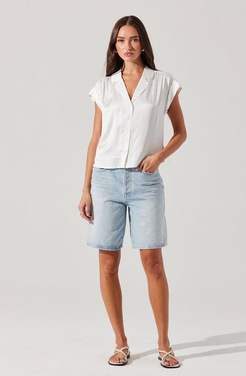 A woman is wearing a white short-sleeved, button-up shirt with a collar and slightly rolled-up sleeves, paired with light wash denim shorts that reach above the knees. She is also wearing white strappy sandals.