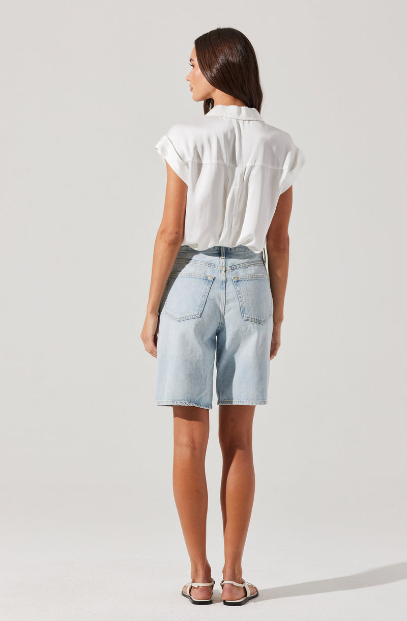 A woman is wearing a white short-sleeved blouse with a collar and a pleat detail at the back, paired with light blue denim shorts that have a high waist and large back pockets. She is also wearing flat sandals.