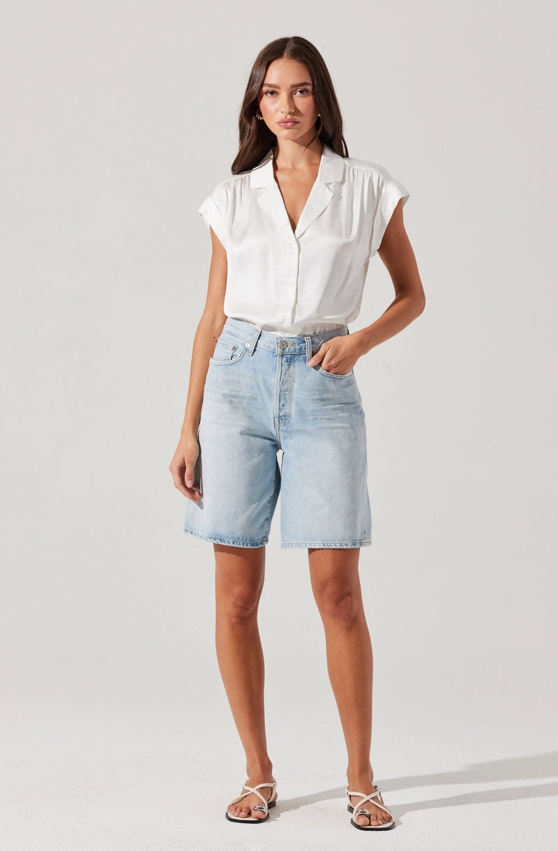 A woman is wearing a white short-sleeved, collared blouse with a V-neck and a loose fit. The blouse features a subtle vertical stripe pattern and is tucked into light-wash denim Bermuda shorts that have a high waist, belt loops, and a relaxed fit. She is also wearing white strappy sandals with a low heel.