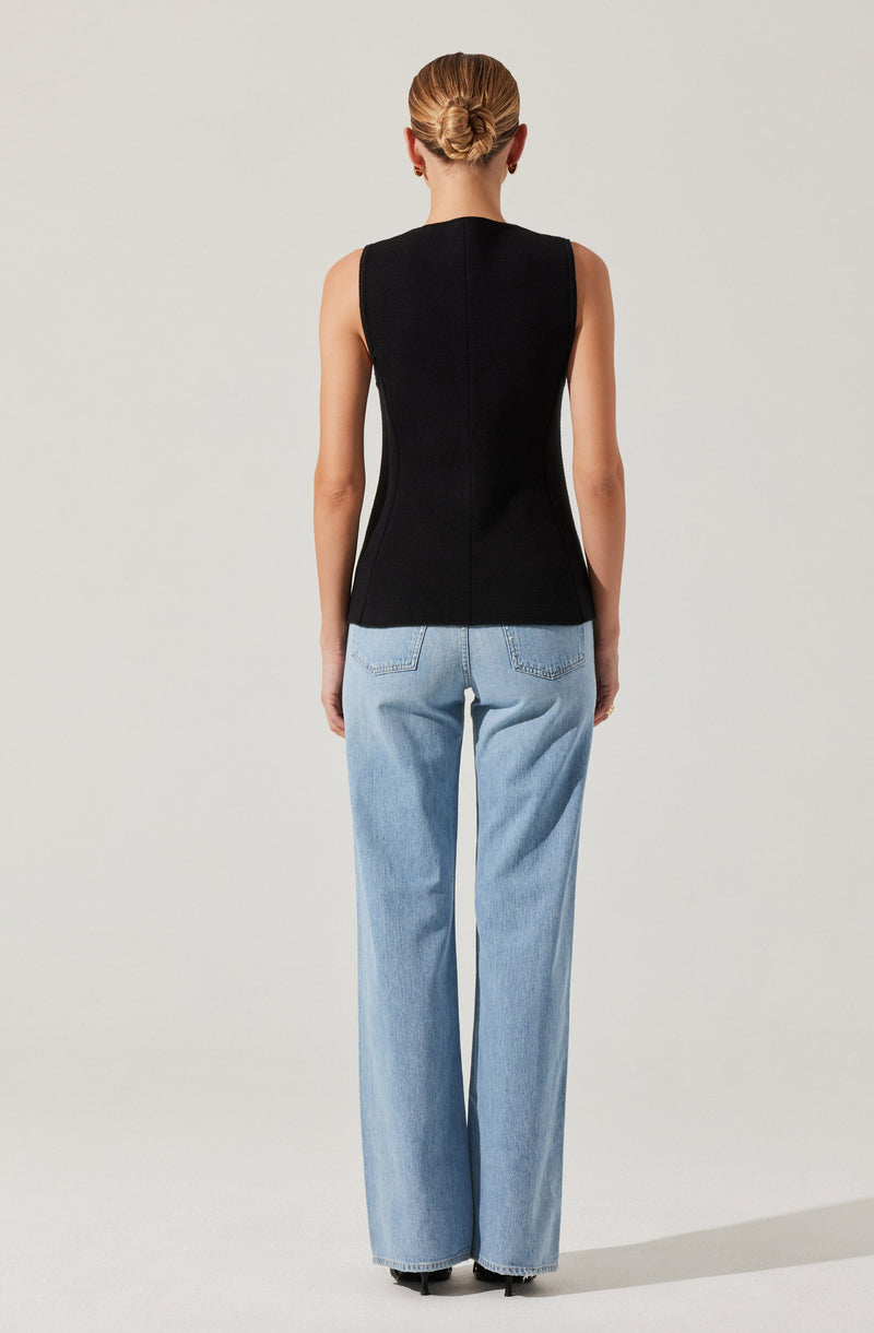 A woman is wearing a sleeveless, fitted black top with a central seam running down the back and light wash, high-waisted, wide-leg jeans. She has her hair styled in a bun and is standing against a plain, light-colored background.