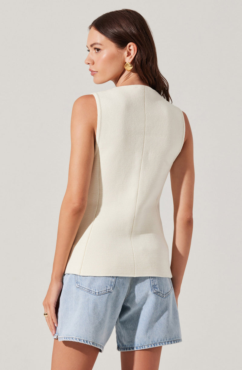 A woman is wearing a sleeveless, cream-colored textured top with a high neckline and visible seam details down the back. She has paired it with light blue denim shorts that have a relaxed fit and rolled-up hems. She is accessorized with large gold earrings.
