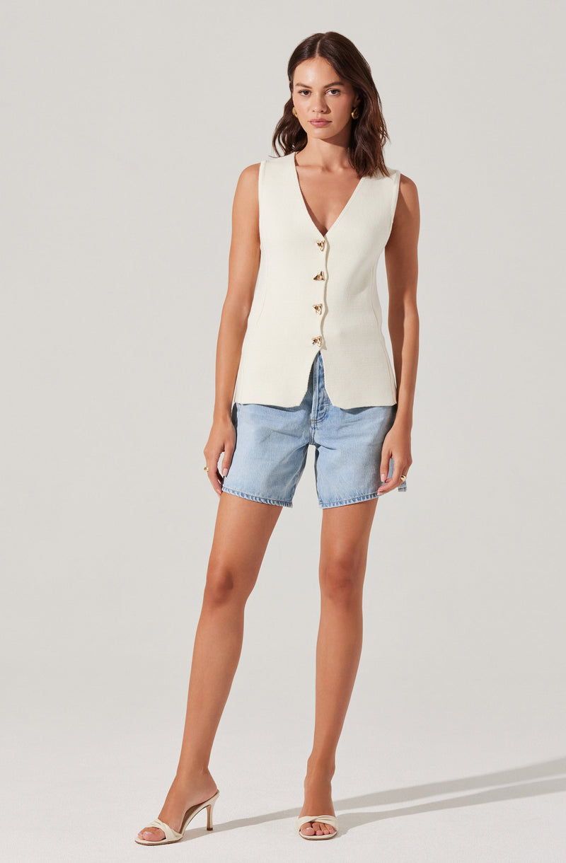 A woman is wearing a sleeveless, cream-colored vest with a V-neckline and gold buttons down the front. She has paired it with light blue denim shorts and beige high-heeled sandals.