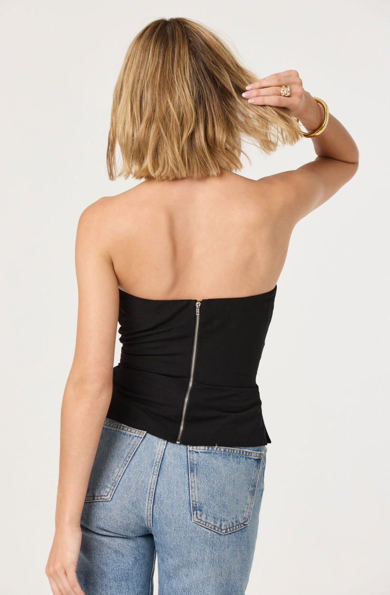A woman is wearing a strapless black top with a silver zipper running down the back and light blue denim jeans. She has shoulder-length blonde hair and is accessorizing with a gold bracelet and a gold ring.