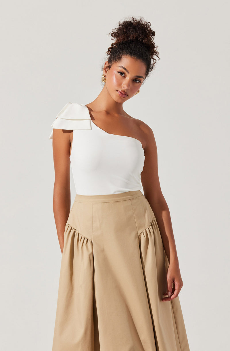 A woman is wearing a white, one-shoulder top with a layered ruffle detail on the shoulder strap. She pairs it with high-waisted, beige wide-leg pants that have a gathered, pleated design at the hips.