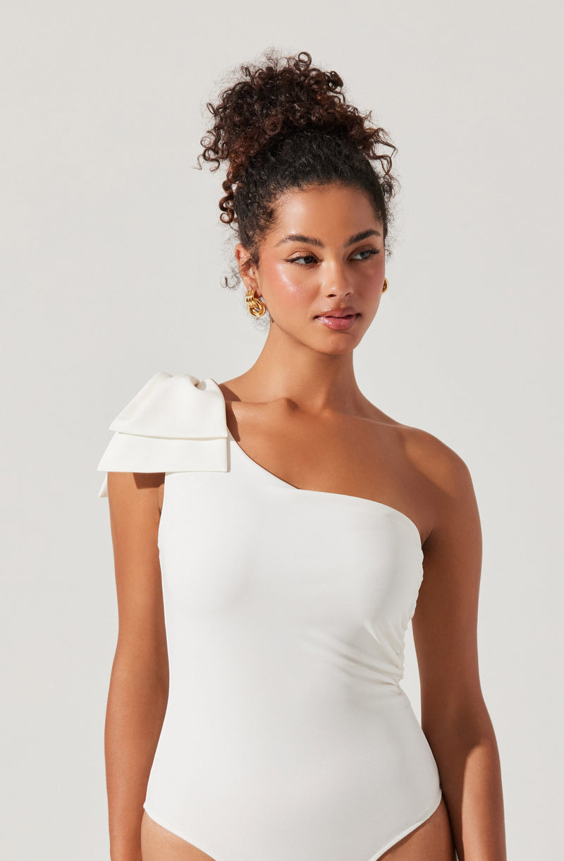 A woman is wearing a white, one-shoulder bodysuit with a large decorative bow on the shoulder strap. The bodysuit has a sleek, fitted design, highlighting her figure.