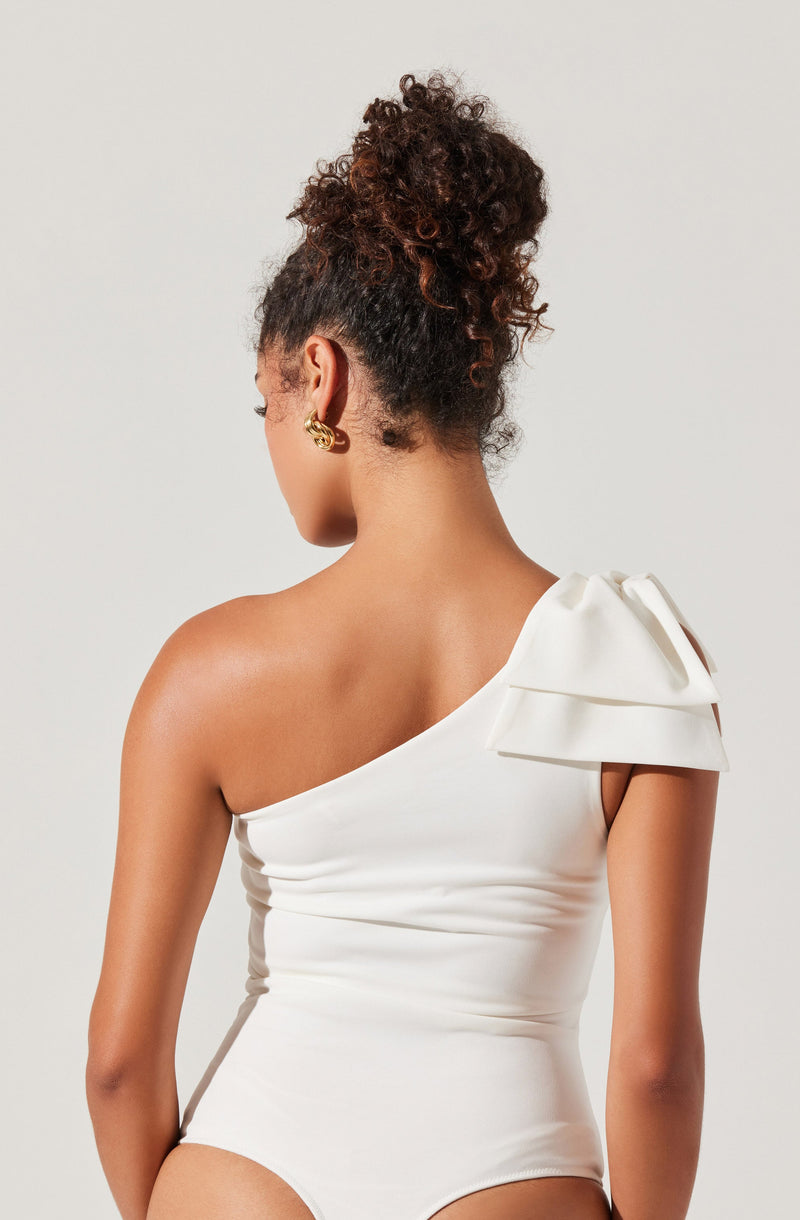 A woman is wearing a white, one-shoulder garment with a large, decorative bow on the shoulder strap. The fabric appears smooth and fitted, emphasizing her back and shoulders. She has curly hair styled in a high bun and is accessorized with gold earrings.